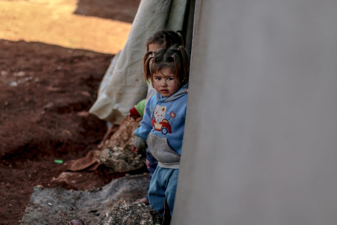 About Children stand near a tent in a refugee camp in Idlib, Syria, reflecting resilience and hardship.